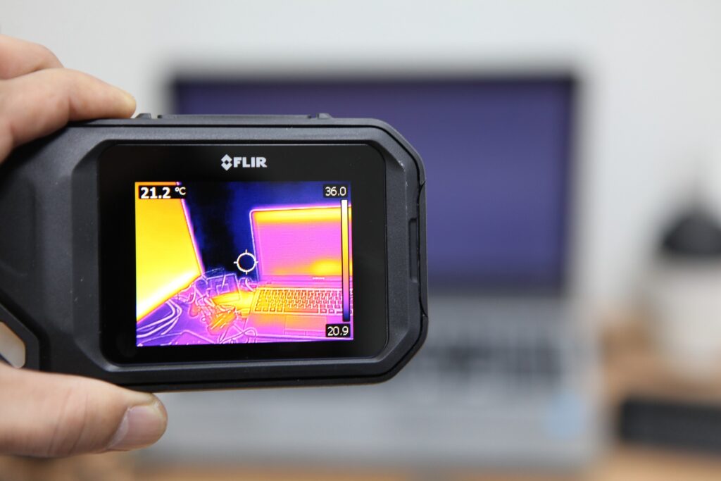 Trzymana w dłoni kamera FLIR z termicznym obrazem komputera na ekranie; w tle widoczny laptop