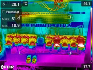 Kolorowy termogram wnętrza rozdzielnicy z przewodami, aparatami elektrycznymi oraz skalą temperatur po prawej